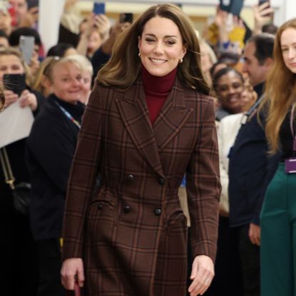 Kate Middleton wearing a plaid coat and red turtleneck smiling walking away from a crowd of staff at a hospital 