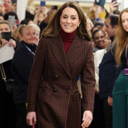 Kate Middleton wearing a plaid coat and red turtleneck smiling walking away from a crowd of staff at a hospital 
