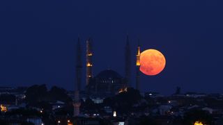 large orange fully illuminated moon rising in the sky behind a large mosque.