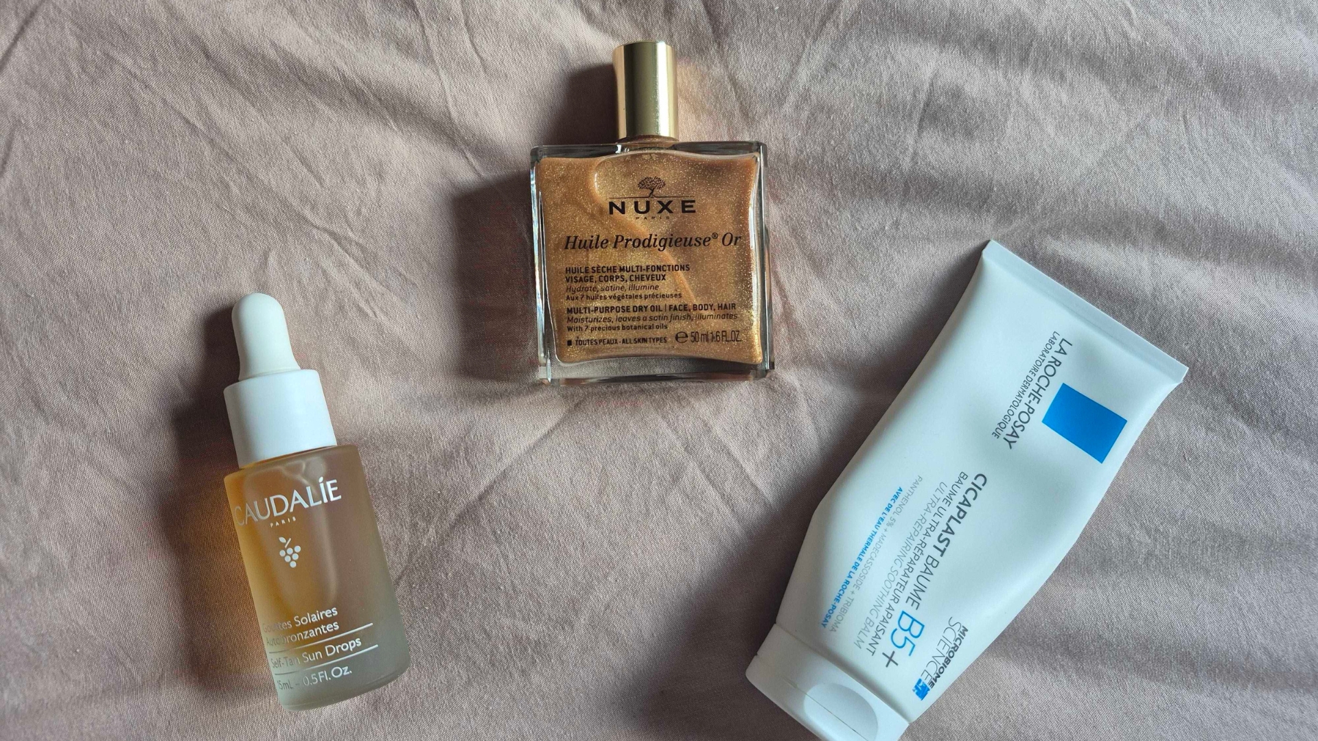 A picture taken by Digital Beauty Writer, Naomi Jamieson, of three French beauty products featured in this round-up including the Caudalie Self Tan Sun Drops, the Nuxe Prodigieuse Shimmering Dry Oil and La Roche-Posay Cicaplast Baume B5, on a pink bedding background