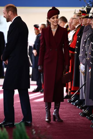 Kate Middleton wearing a maroon coat and hat walking a long a row of uniformed men with Prince William standing to the side