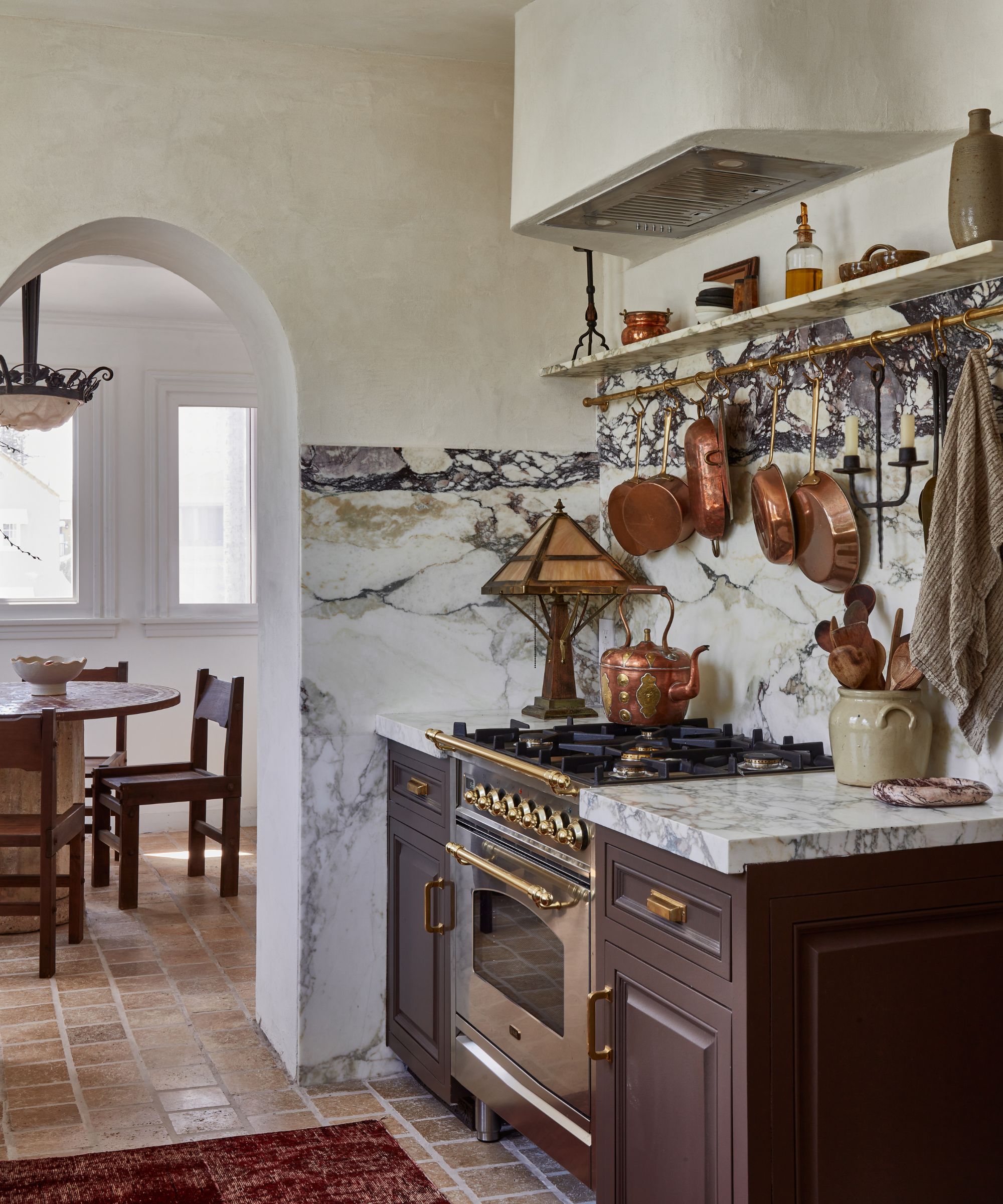 A Vintage kitchen with brown cabinets, a marble countersplash, a brass pot rack on the wall, and a range cooker topped with a vintage copper teapot