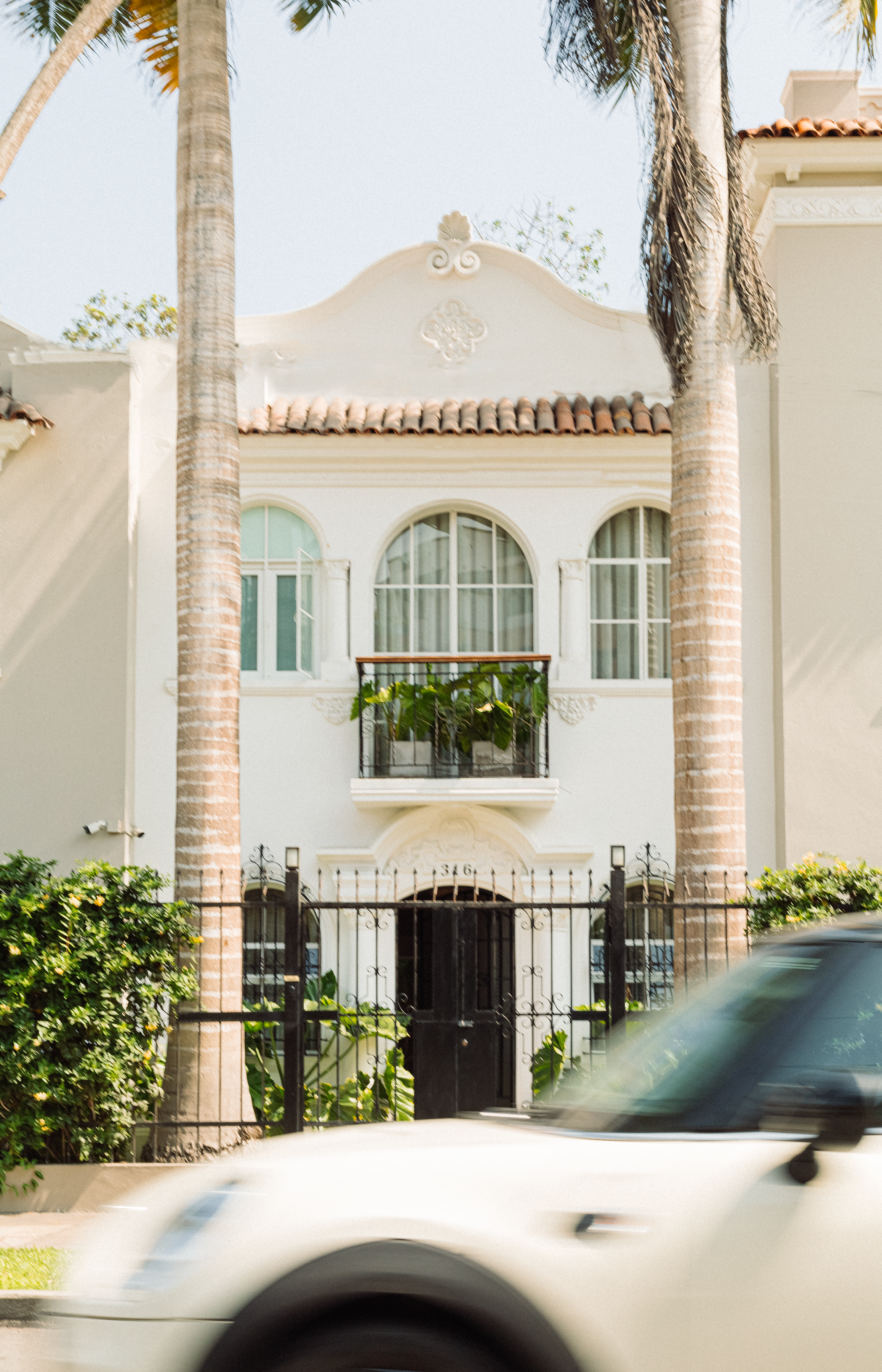A blurred beige car in front of an historical building with plants-decorated balconies and an equally lush outdoor patio, captured in the sunshine.