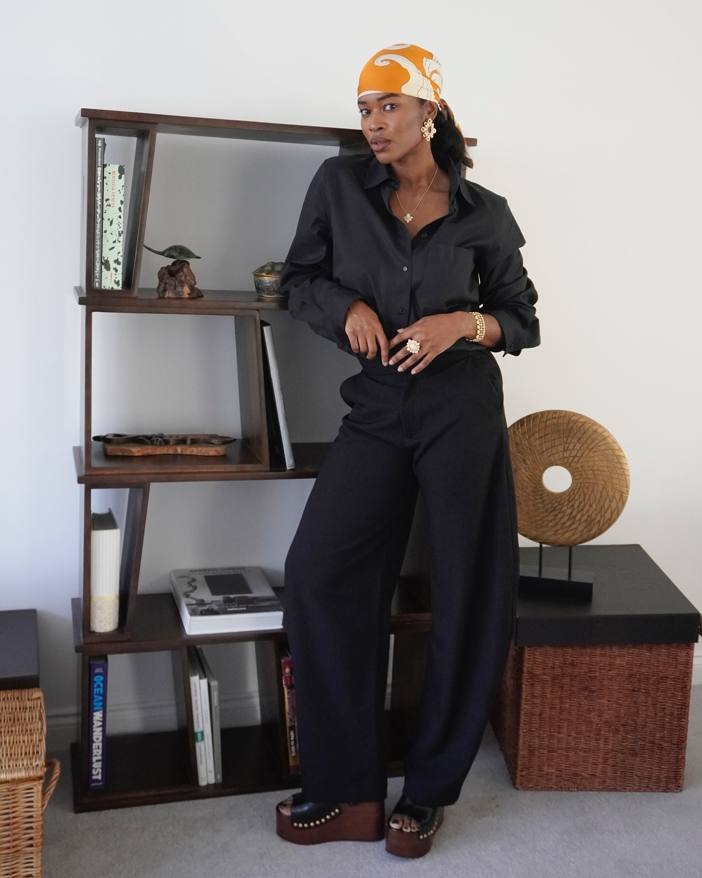 Woman wears a black button down and trousers with a marigold printed silk head scarf.