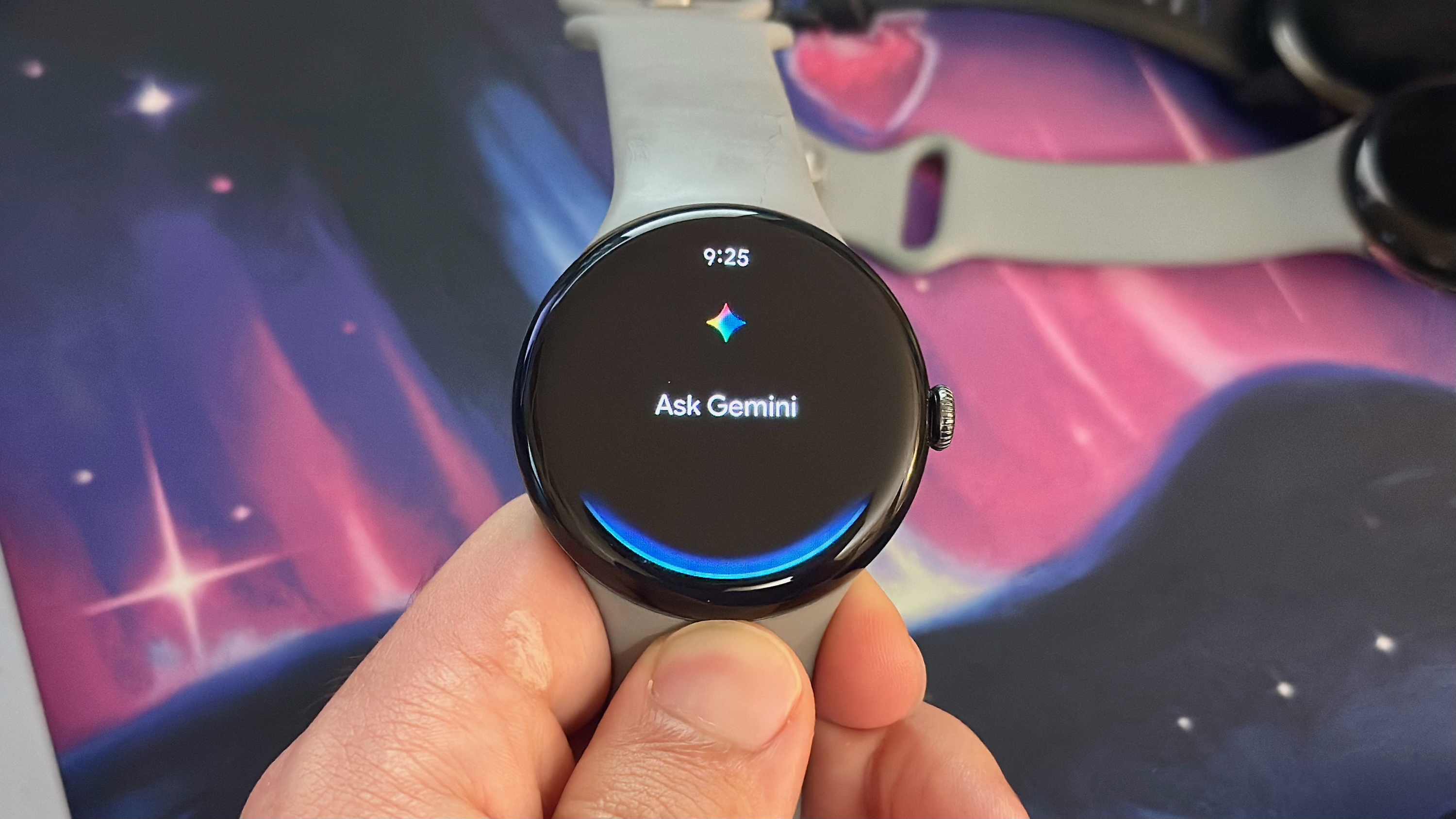The Google Pixel Watch 3 held in hand above a desk, the words &quot;Ask Gemini&quot; and a listening indicator visible on the display.