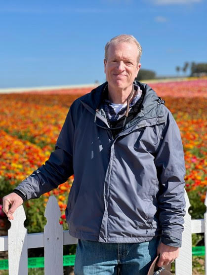 Man stood against white fence in blue jacket, with red flowers growing in a field in the background