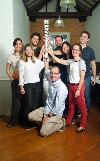 Smiling team of eight male and female designers, grouped together holding up the finished Olympic torch, brick and white walls in the background, wooden floor, dark wood beam and white ceiling, two opaque lit ceiling lights, white framed window on the back wall with view of nearby tree branches