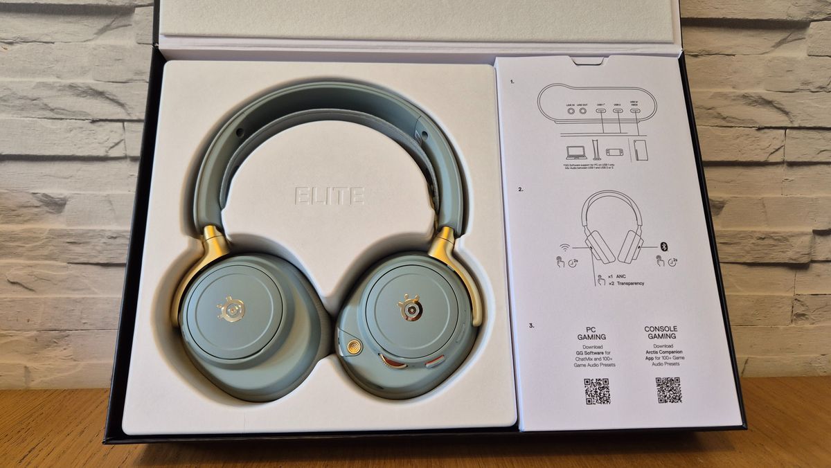 The SteelSeries Arctis Nova Elite headset in front of a white brick background and on a wooden surface