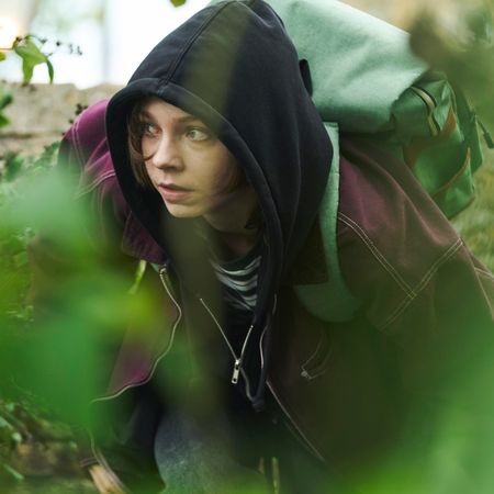 emma myers as pip wearing a hoodie purple jacket and backpack as she hides in grass in a good girl's guide to murder season 3