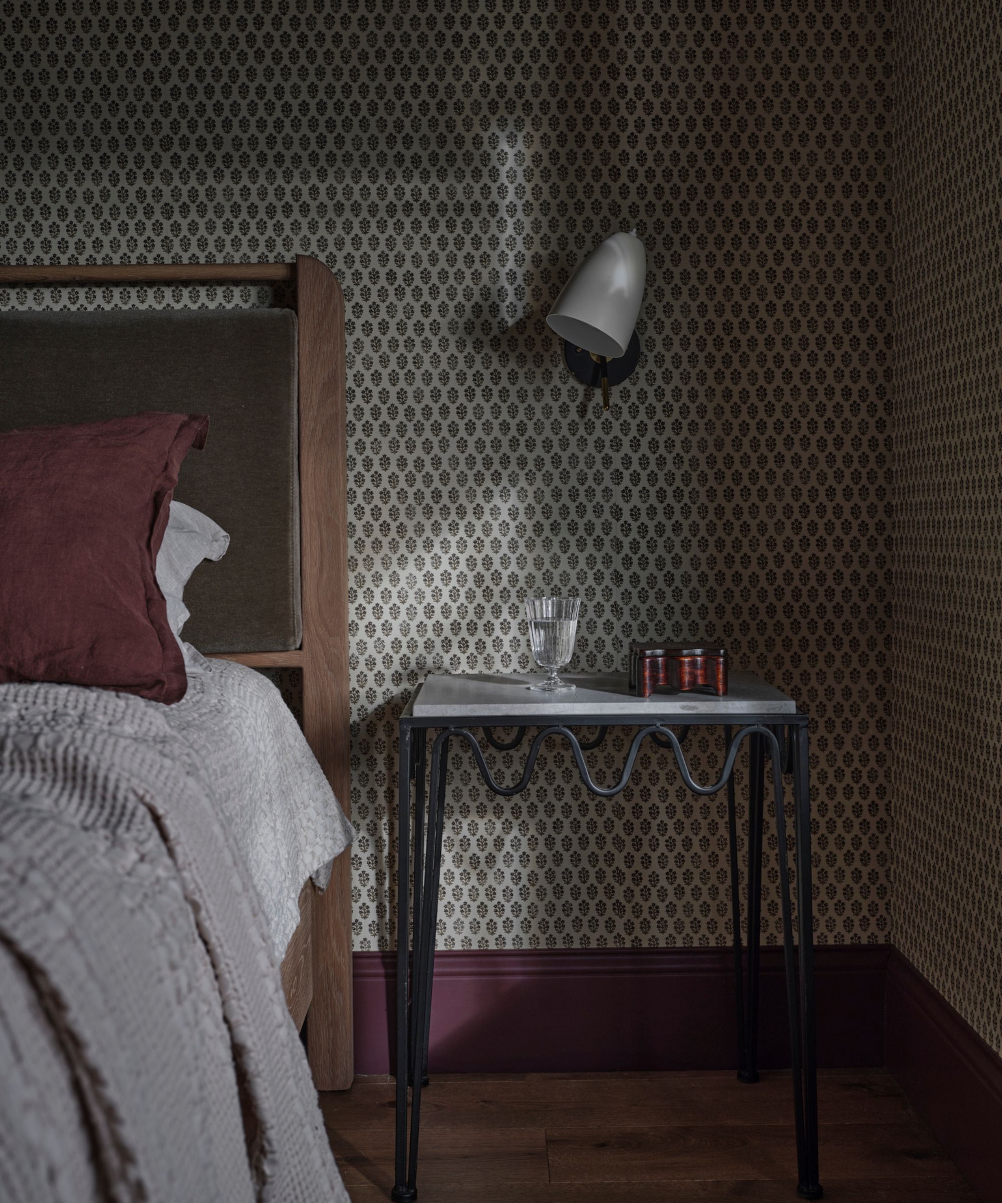 a small cottage bedroom with ditsy wallpaper and purple skirting boards, a wooden bed with green velvet headboard and a wavy metal nightstand