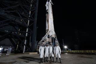 four people in white spacesuits with the visors up pose and smile in front of a white rocket upright on a launchpad