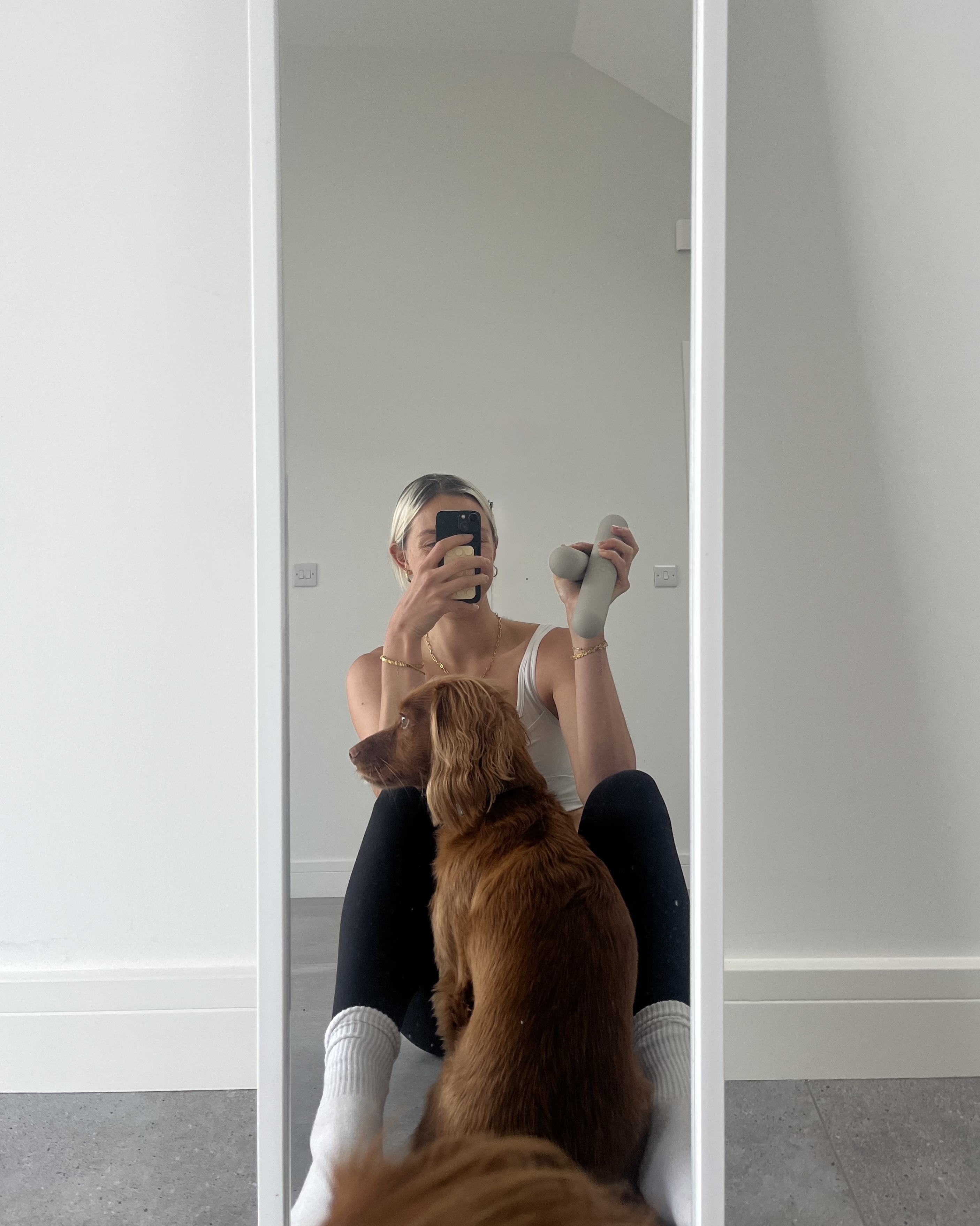 A woman takes a mirror selfie holding small hand weights with her pet dog