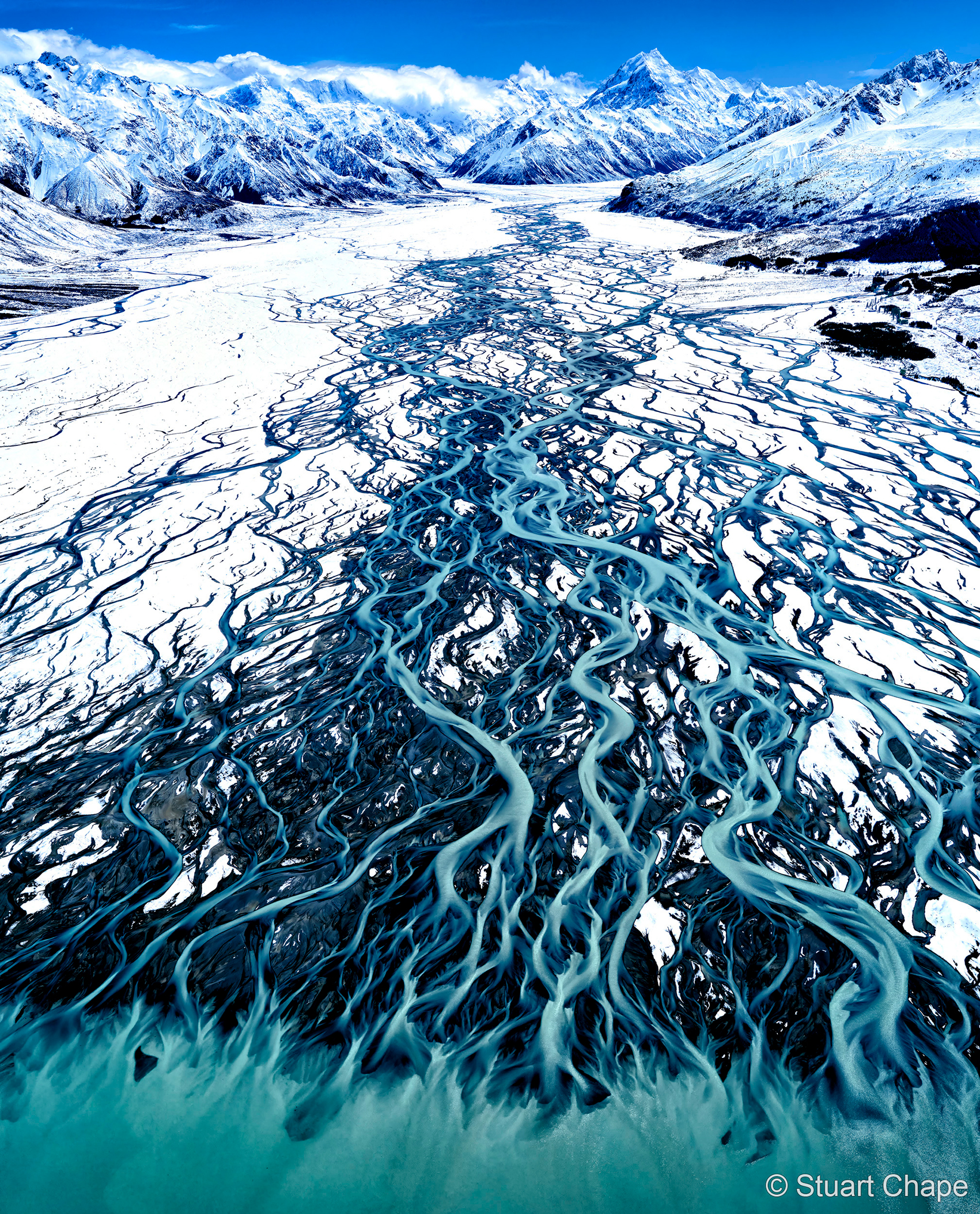 Aerial view of a glacial river meandering through a snowy landscape, surrounded by towering mountains and blue sky