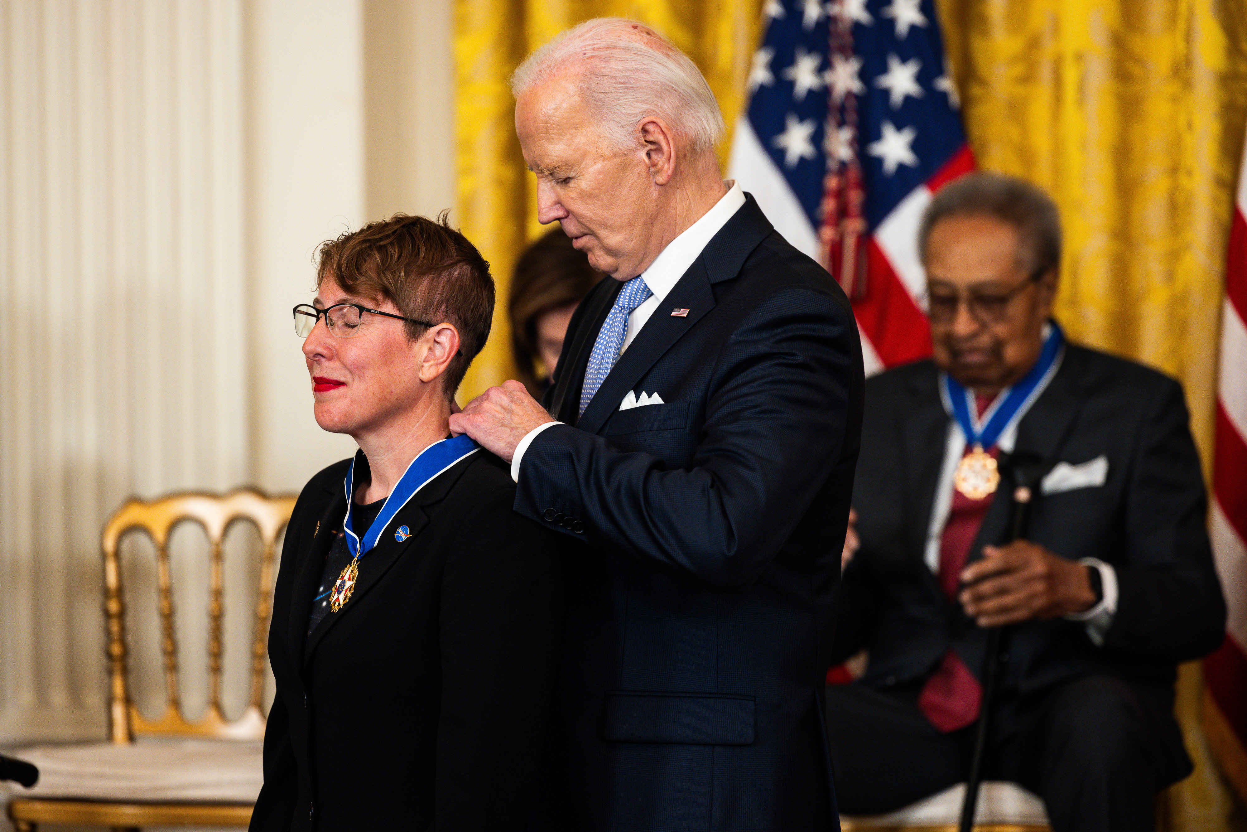 una mujer que lleva un pin de la NASA sonríe con lápiz labial rojo mientras un anciano con traje detrás de ella le pone el collar de una medalla que cuelga alrededor de su cuello.