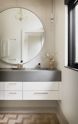 A light off white bathoom with a grey top vanity unit and oversized round wall mounted mirror