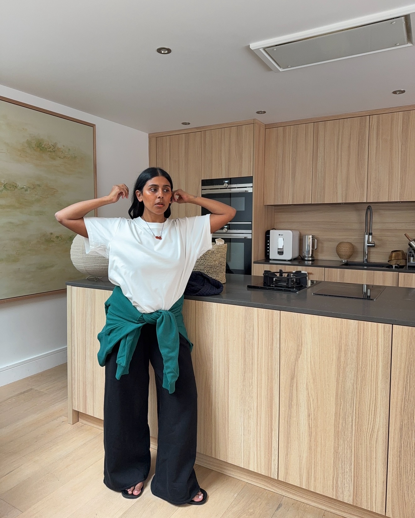@monikh wears a pair of black wide-leg trousers with a white T-shirt and a teal jumper around her waist