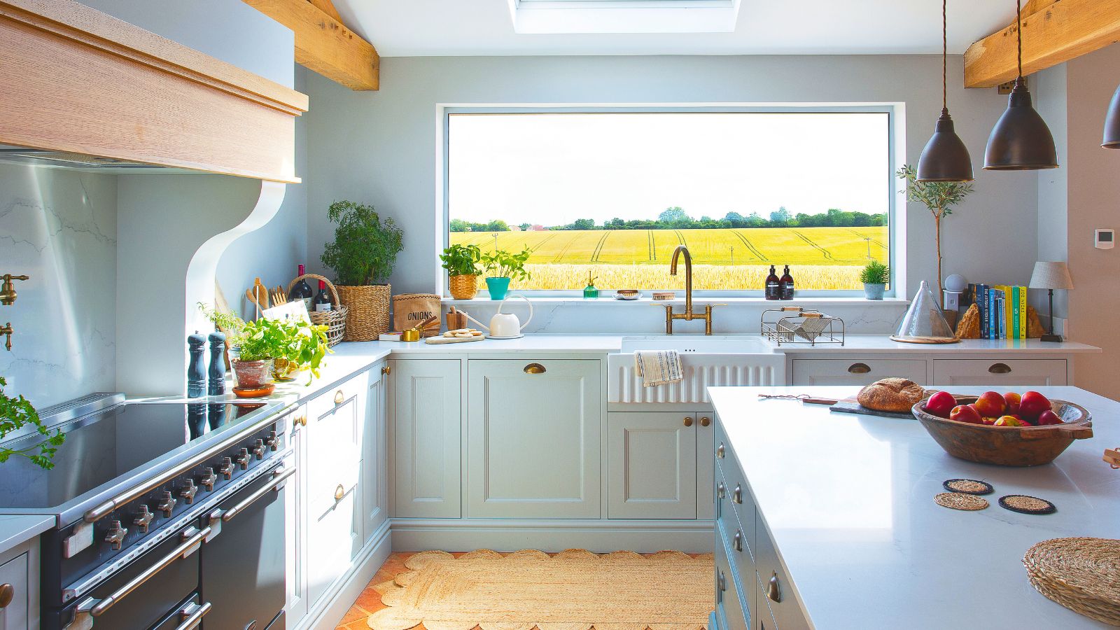 A light blue kitchen with white counters, wooden beams above a large range stove. A large window behind the kitchen sink framing green fields. 