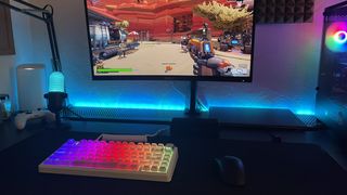 A white RGB gaming keyboard on a black desk with a monitor overhead showing Roboquest