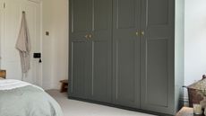 Image of a sage green wardrobe in a white room with off-white carpeting, a blue rug, and a bed with sage green bedding.