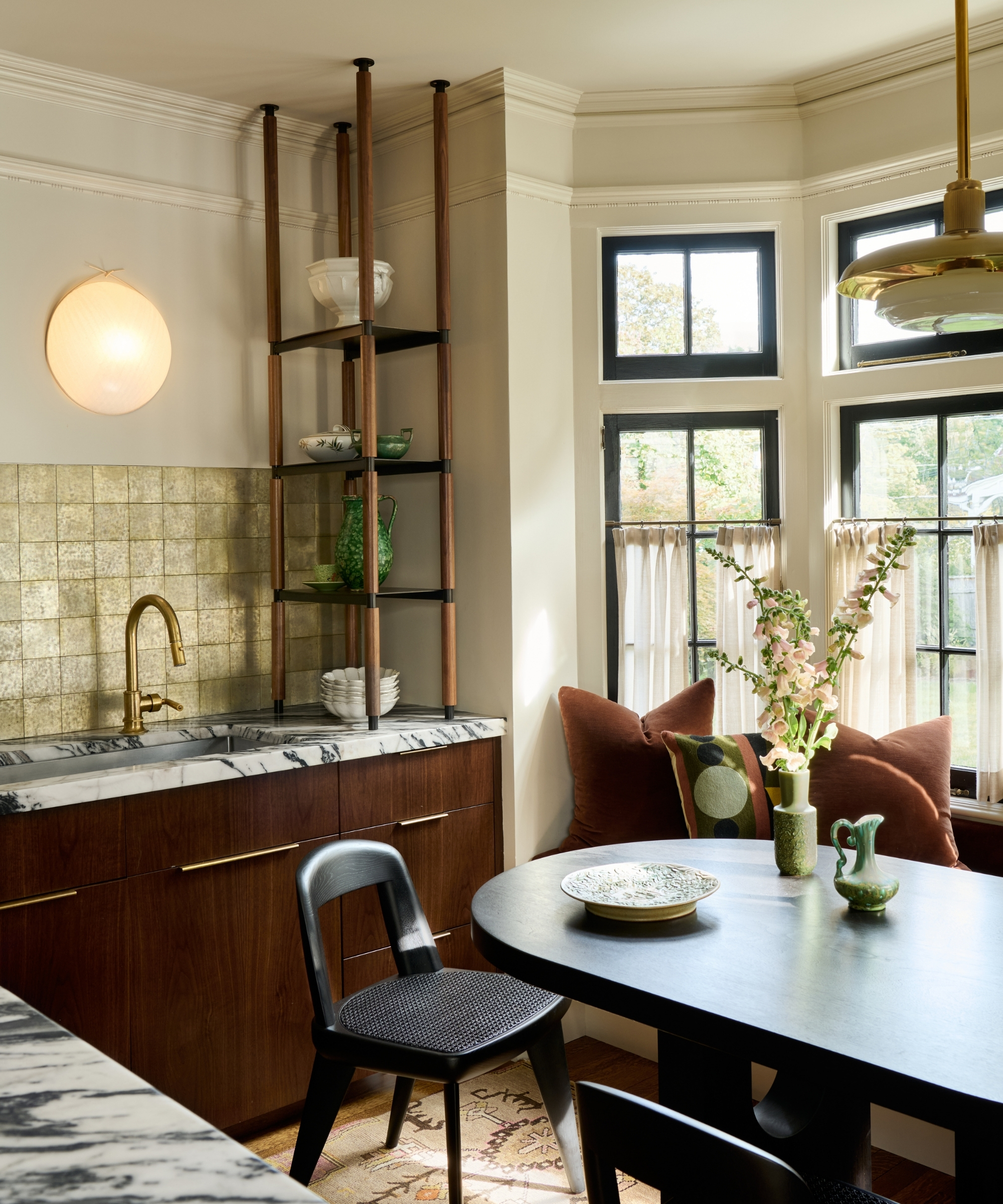 The corner of a kitchen with dark wood cabinets, marble countertops, and a bay window used for a banquette seat with an oval black table