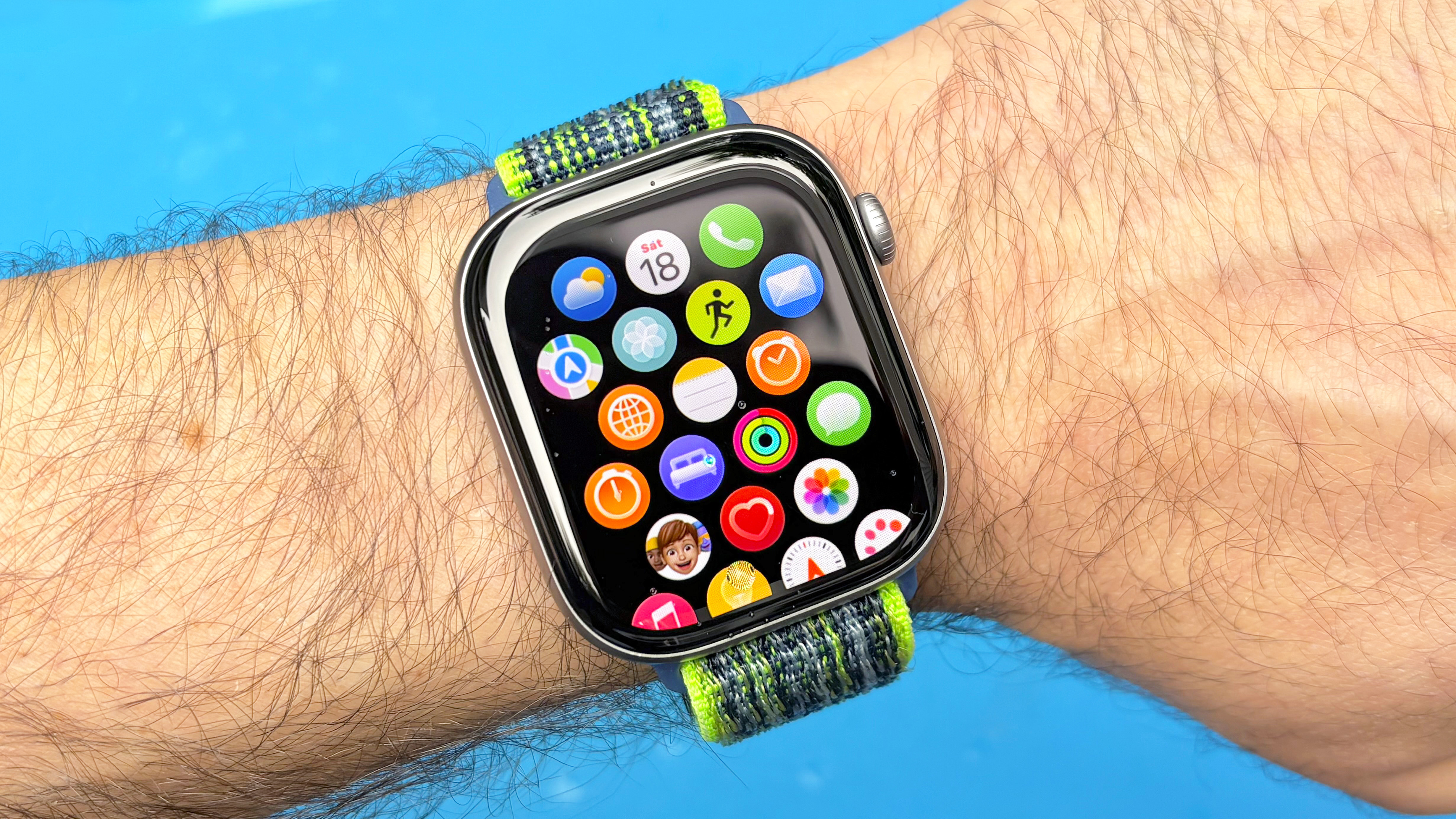 Close-up of the Apple Watch 11 on a user's wrist against a blue background with the app icon grid displayed on the screen
