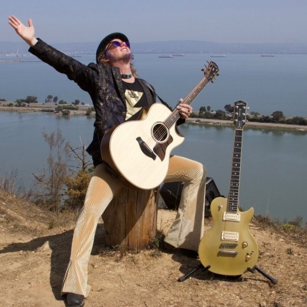 Jimmy Leslie holding an acoustic guitar while sitting on a tree stump