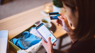 A woman holding a credit or debit card in one hand and carrying out an online purchase on her phone with the other.