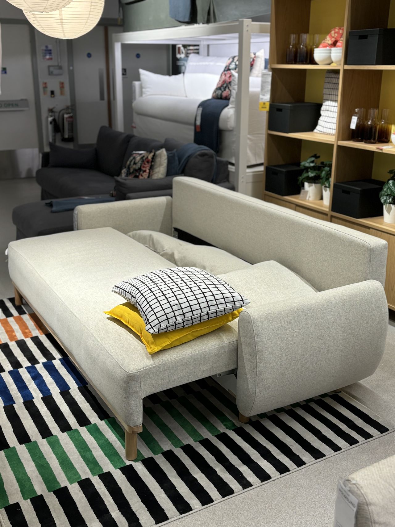 a tan sleeper sofa in a showroom, fully open, with a yellow and blue/white pillow on top.
