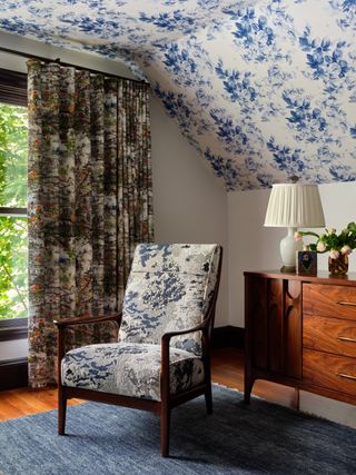 corner of attic bedroom with patterned wallpaper on ceiling, a patterned armchair, patterned curtains, blue floor rug, wooden console with table lamp, picture and vase of flowers