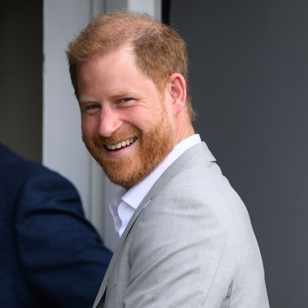Prince Harry wears a gray suit jacket, a white shirt, and dark slacks and has red hair as he visits the UK in September 2025