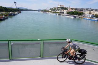 Image shows Anna cycling over the Old Bridge looking out to the Bratislava castle and UFO tower