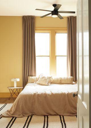 yellow bedroom with taupe drapes, fan, stone bedding, textured bed pillows, side table, modern white lamp, stripe rug, hardwood floor