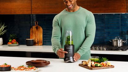 The Nutribullet Ultra making a green smoothie on a countertop with vegetables on a chopping board to the left and a man with a green jumper behind the Nutribullet Ultra 