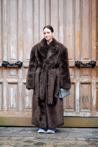 A woman wears a long brown fur coat with a tie belt, jeans, sneakers, and a gray hermes bag.