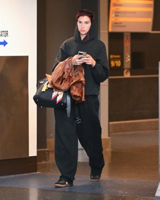 Dua Lipa wearing a black sweatsuit, black PUMA sneakers, and a black Hermès Birkin bag with bag charms at JFK Airport.