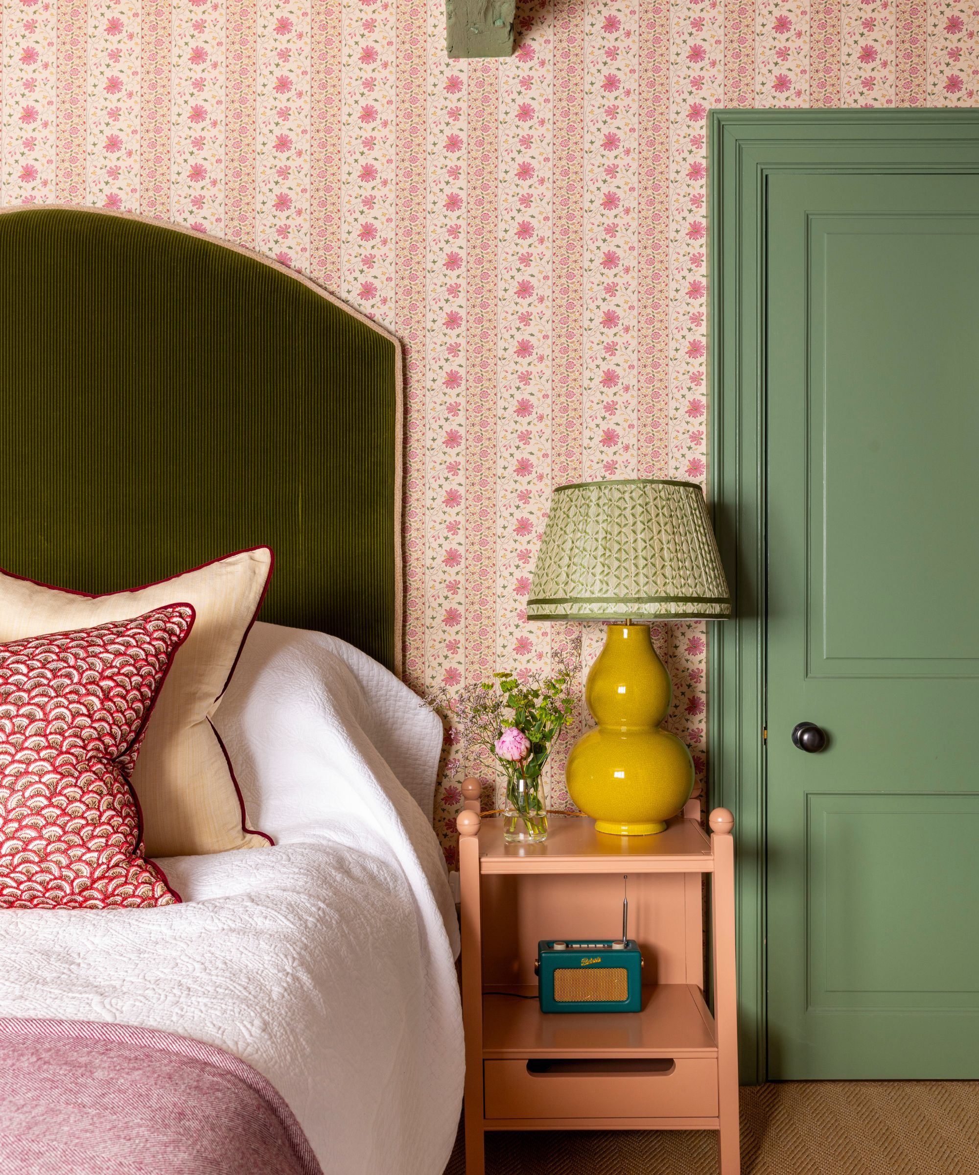 The corner of a traditional bedroom with pink wallpaper, a green door, an olive green headboard and pink cushions.