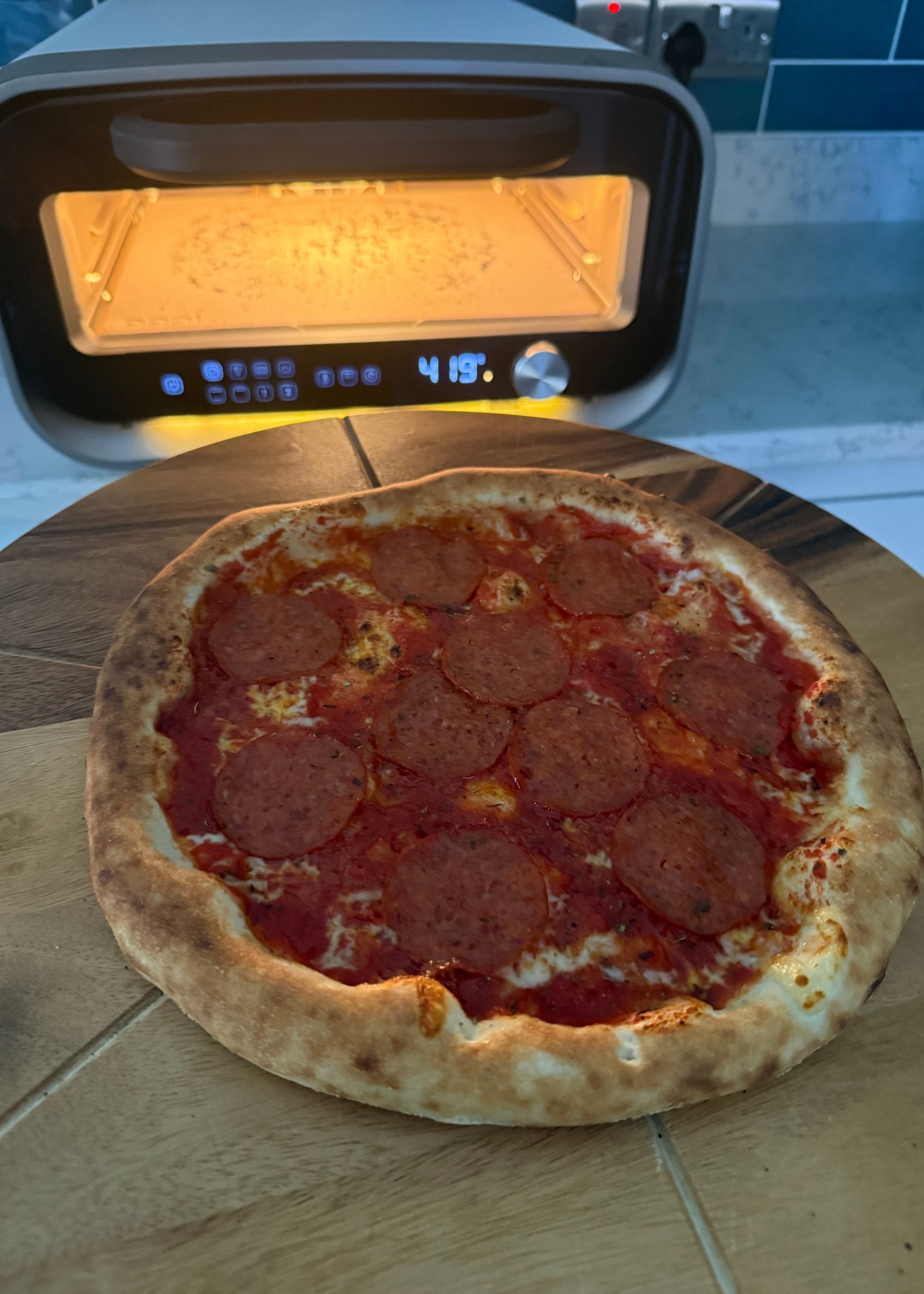 A close up of a pizza on a wooden chopping board with grooves next to Ooni's volt 2 that was used to cook it on a kitchen countertop, still with the oven light on post cooking