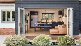 external image of house with dark wood cladding and pale green bifold doors opened showing living room