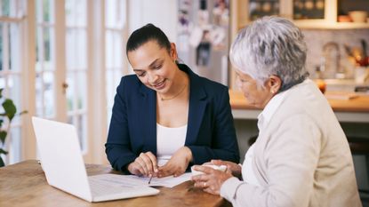 A retiree and a financial adviser sit at a table going over paperwork with a financial adviser.