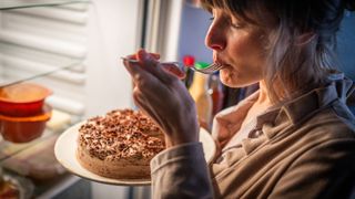 A blonde woman eats a chocolate gateau from the fridge at night before bed