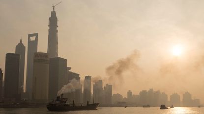 A smog-filled cityscape on the edge of a body of water is seen under a clouded over sun