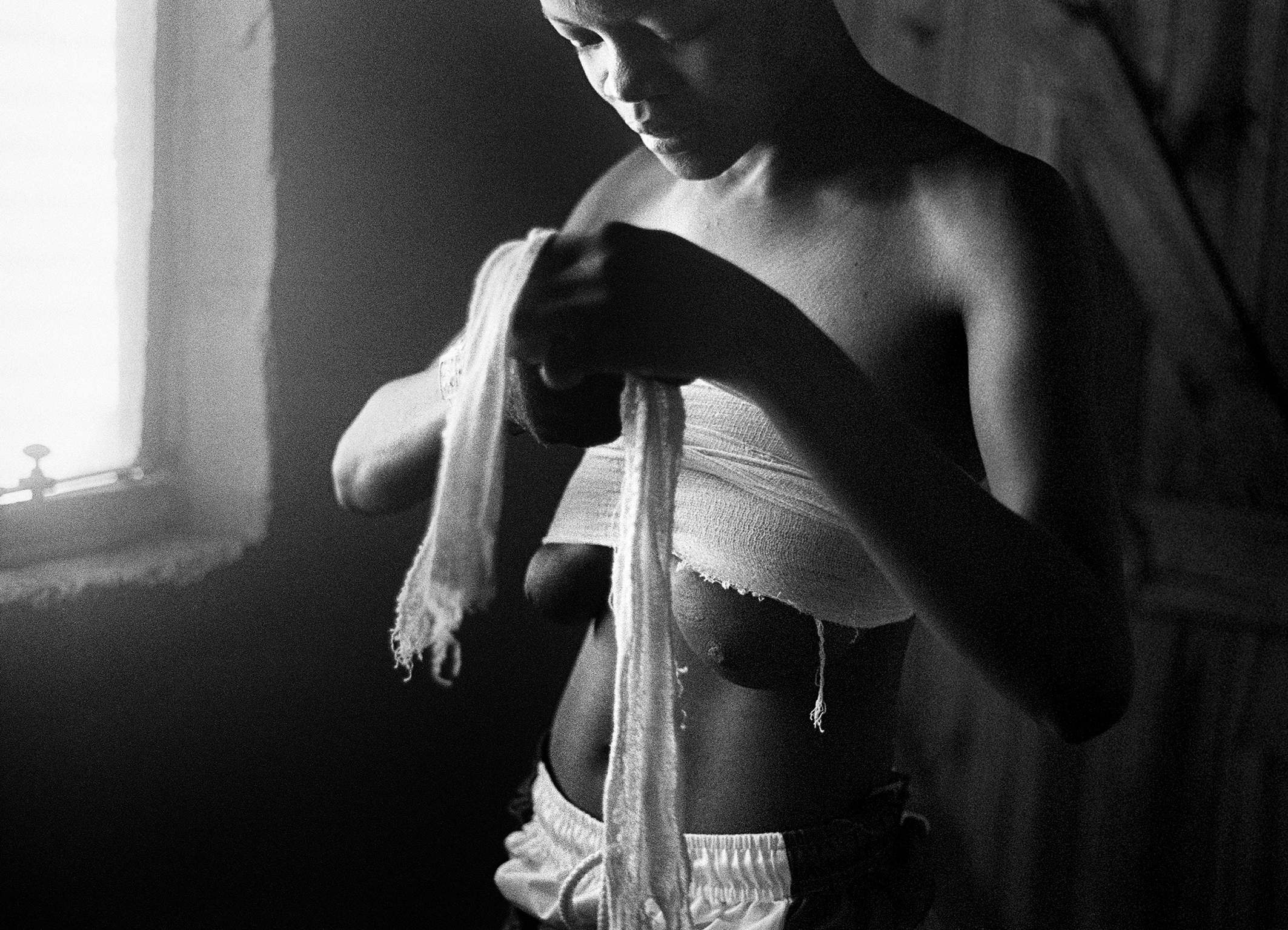 A person stands near a softly lit window, focused on wrapping a cloth around their chest. It's a black and white image