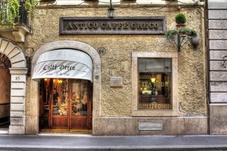 The outdoors of an historical cafe features a white curtain with belle époque font reading "Caffe Greco" in black on a wooden door showing the inside of a warmly lit room. Next to it is a window reading "TEA ROOM" and "CAFFE 1760 GRECO" in golden, upper-cases characters.