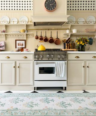 A kitchen with butter yellow walls with shelves with plates on, matching cabinets, a white stove with a yellow kettle on top, and a floor with a green and white floral rug on top