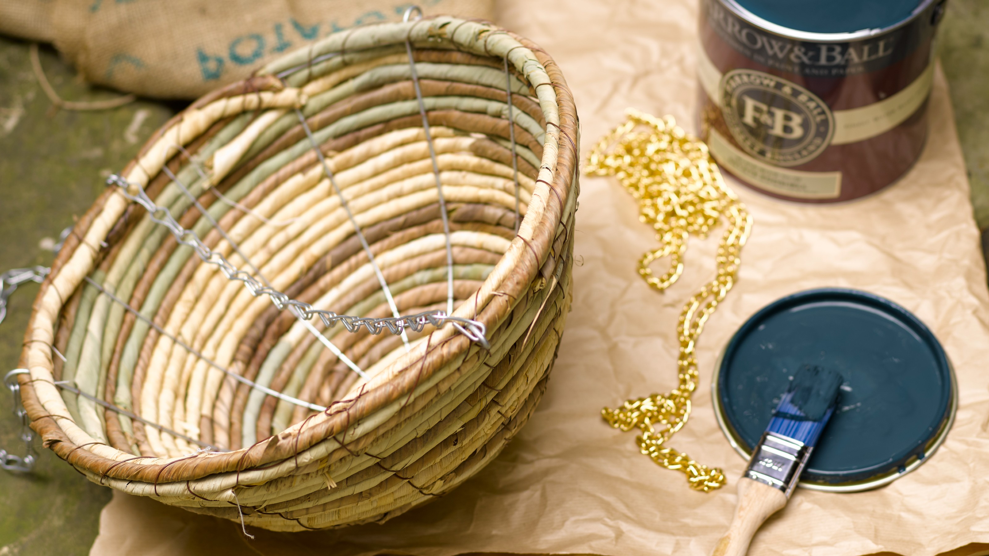 An open tin of blue Farrow & Ball paint with a brush and a rattan hanging planter