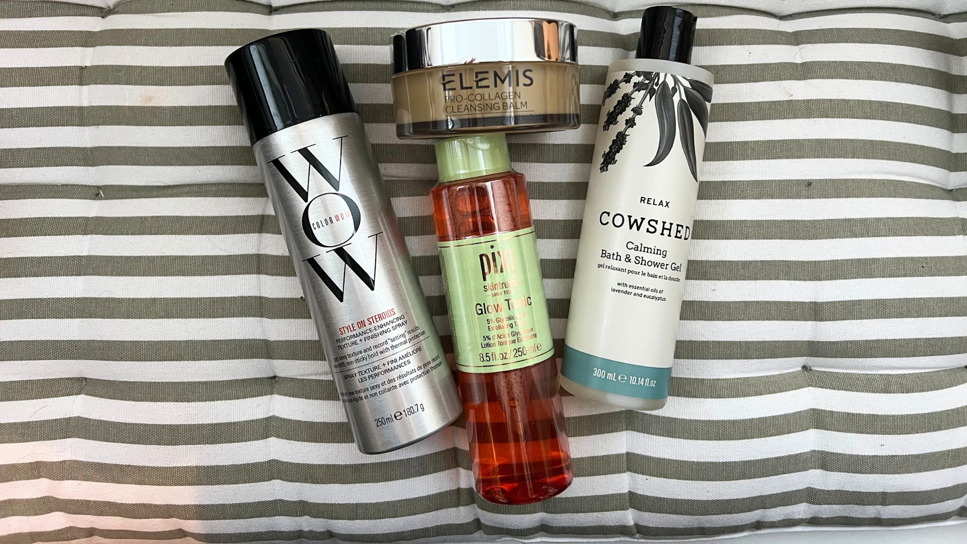 a selection of lookfantastic products pictured on a stripy cushion, pixi glow tonic color wow style on steroids spray, cowshed relax bath and shower gel, elemis cleansing balm