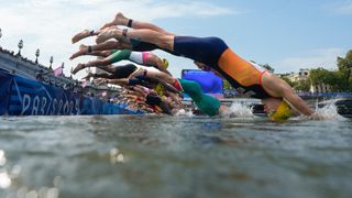 A row of swimmers dives into the Seine river with a banner reading "paris 2024" visible in the background