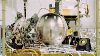 A complex piece of space hardware, featuring a large, shiny metal sphere, is surrounded by a dense tangle of cables, wires, and electronic components in a laboratory.