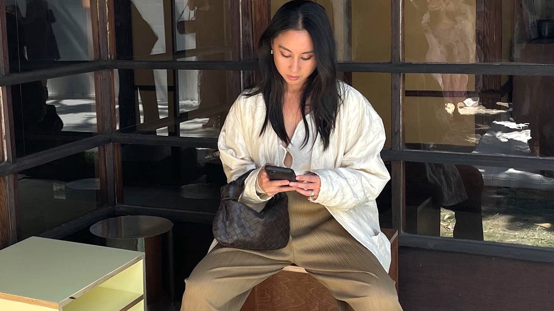 style influencer Jordan Risa sitting outside the Schindler House in West Hollywood wearing a white keyhole tie-front top, a woven brown bag, and pliss&eacute; tan pants
