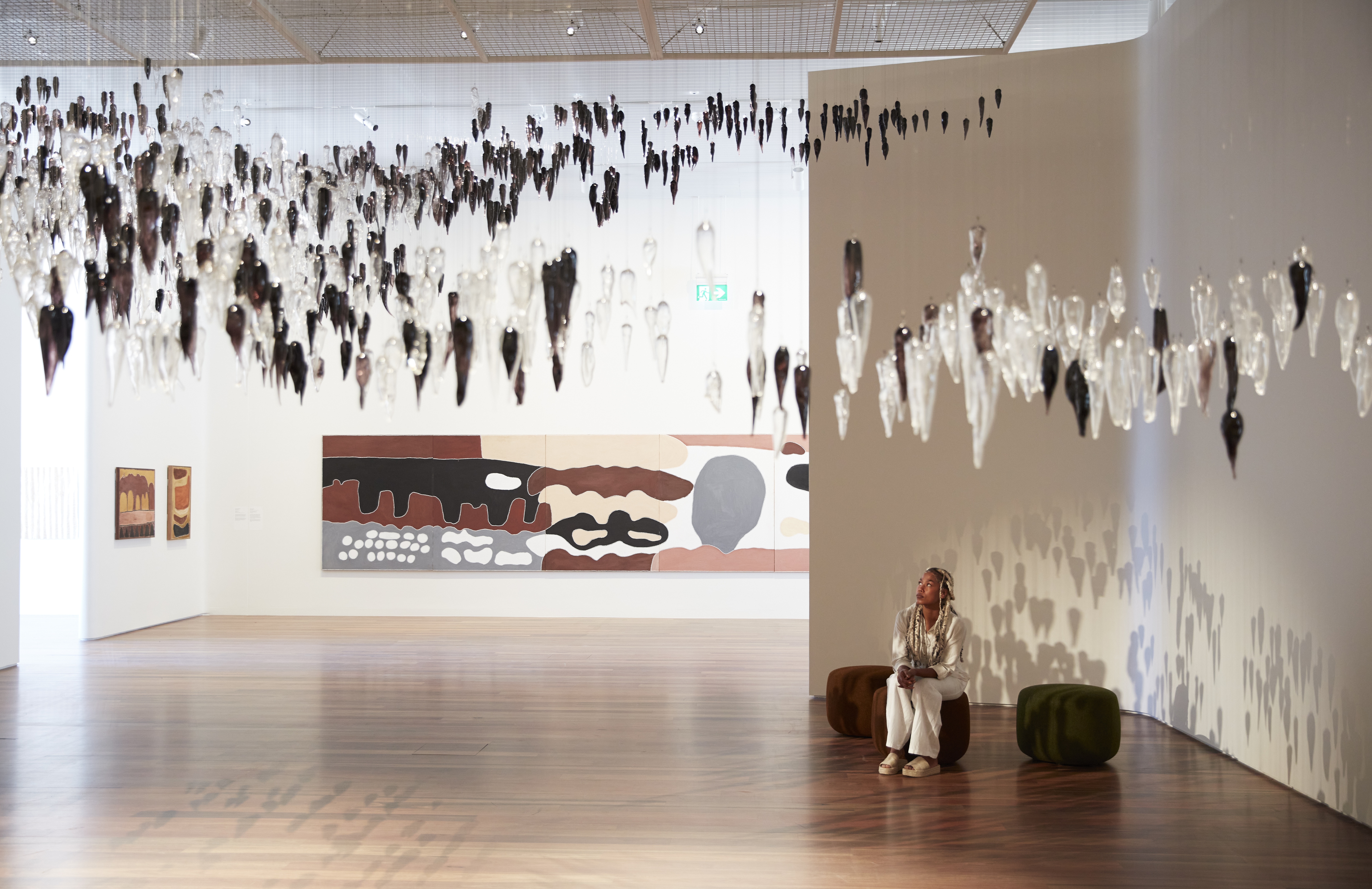 A museum gallery is filled with drop-like floating sculptures, earthy large-scale paintings, and abstract, comfy stools a woman is currently sitting on.